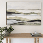 Framed abstract landscape painting with layered rolling hills in soft beige, green, and gray tones above a wooden console table with a potted plant and a small bird figurine on books.
